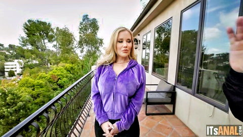 Blonde babe in purple velour hoodie stands on balcony with man outdoors