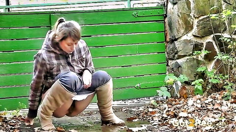 Cute brunette in plaid shirt squatting to pee outdoors behind fence