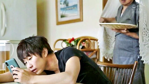 Young Asian guy intently checks phone while maid serves food in home kitchen