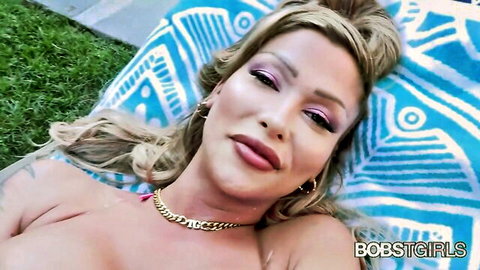 Busty blonde with big tits lying topless outdoors on blue blanket smiling seductively.