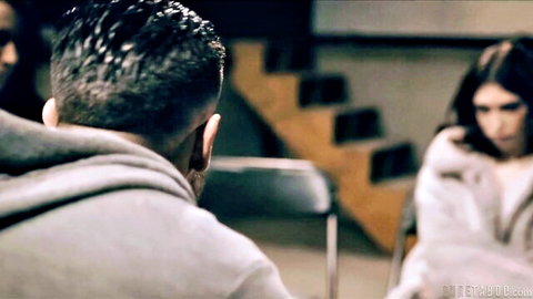 Skinny brunette in fluffy white robe sits facing muscular man in gray hoodie in dimly lit room