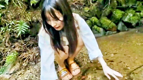 Asian woman in white top squats by forest stream, casual outdoor pose