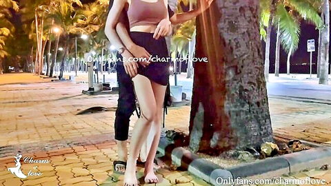 Slim Asian woman in pink bralette and shorts hugs man by palm tree at night outdoors