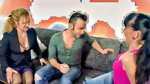Two sexy women in revealing outfits seduce a man on couch in hot threesome preview