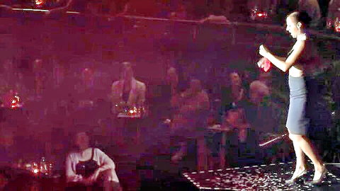 Brunette MILF in short skirt stripteasing on stage holding red bra amid candlelit audience