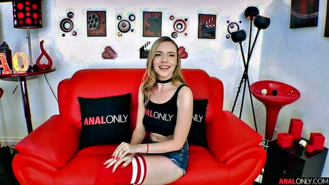 Smiling young blonde in tight ANAL ONLY tank top and denim shorts teasing on red couch