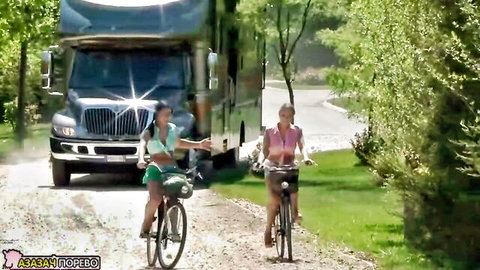 Two busty blondes in skimpy tops and shorts bike outdoors near RV on dirt path