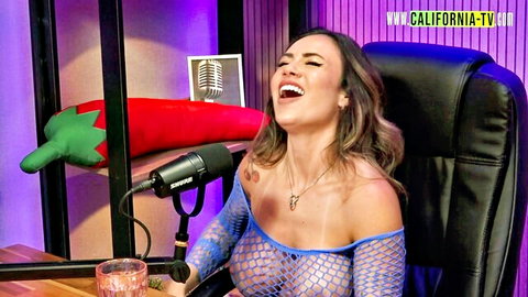 Busty Latina laughs with huge tits exposed through sheer blue fishnet top on TV show