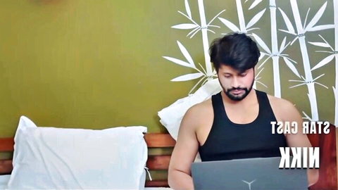 Bearded Indian man in black tank top sits on bed using laptop in green bedroom