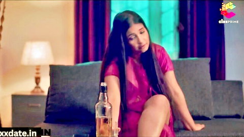 Seductive Indian woman in sheer pink dress spreads legs on couch holding whiskey bottle teasing