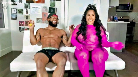 Asian babe in sheer pink mesh bodysuit flaunts big tits and ass beside muscular Black stud in bulging briefs holding yellow butt plug