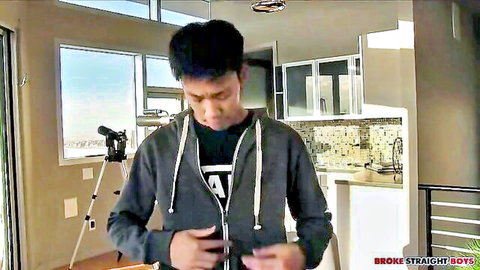 Cute Asian guy in gray hoodie stands in kitchen gripping zipper teasing cock bulge