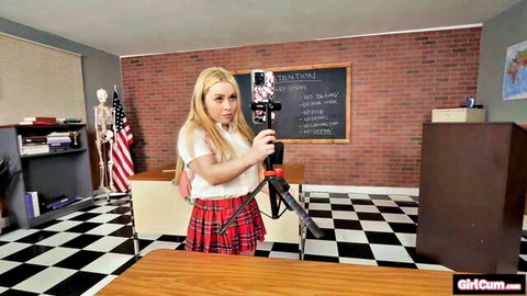 Blonde schoolgirl in plaid skirt holding camera tripod in detention