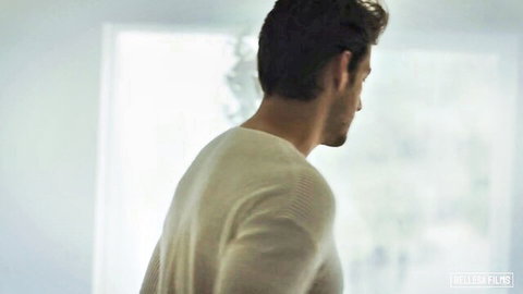 Handsome muscular man in white sweater stands by sunlit window in Brazzers scene