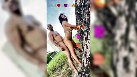 Nude Asian woman with tits out holds orange against tree as naked black man poses behind her outdoors