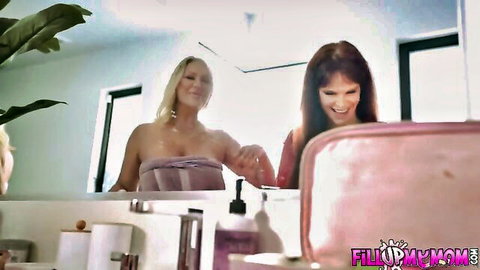 Blonde MILF in towel smiles seductively with brunette in steamy bathroom mirror