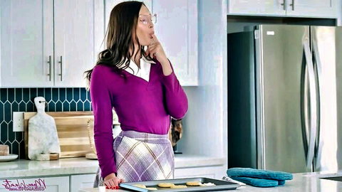 Brunette housewife in purple sweater and plaid skirt teases with finger to pursed lips holding cookies in kitchen