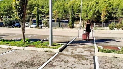 Sexy brunette in short black mini dress walking with red suitcase in outdoor parking lot