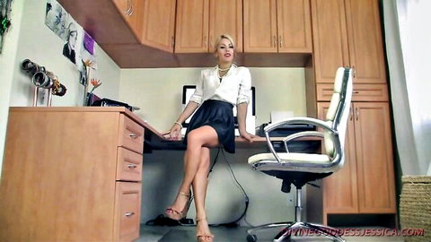 Blonde femdom boss in white blouse and short black skirt sits teasingly on office desk with legs crossed in heels