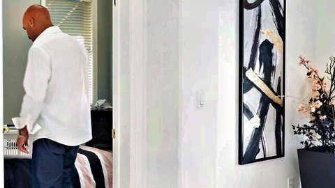 Bald black man in white shirt standing in bedroom doorway adjusting pants
