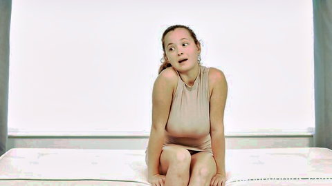 Redhead babe in tight beige dress sits on bed with surprised open-mouth expression