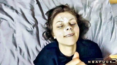 Young brunette teen smiles with thick messy cumshot covering her face and eyes on bed