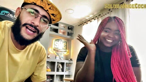 Ebony babe with pink braids smiles and waves in interview with bearded guy in yellow hat