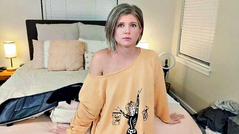 Short-haired gray MILF in off-shoulder orange sweater sits on bed looking at POV camera