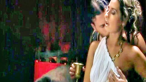 Busty blonde in sheer toga kisses brunette's neck passionately holding golden goblet in fantasy scene
