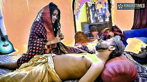 South Asian woman in sheer black saree bends over bare-chested older Indian man lying on bed in steamy taboo scene