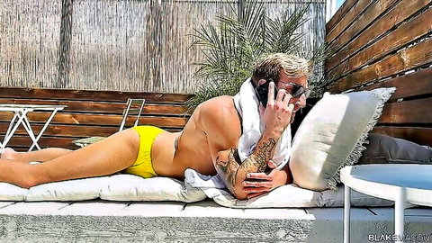 Muscular tattooed hunk in tight yellow speedo bulge lounging outdoors on phone call