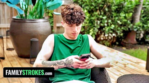 Ethan Seeks in green tank top, curly hair, tattooed arm, checking phone outdoors