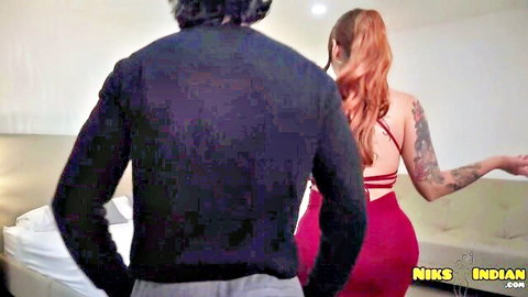 Curvy tattooed redhead with huge tits and big ass in skimpy red backless dress stands as man approaches from behind