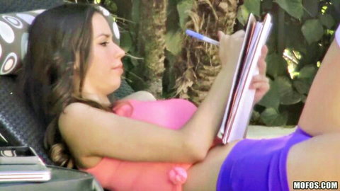 Brunette teen in tight pink tank top and purple shorts lounging poolside writing with pen
