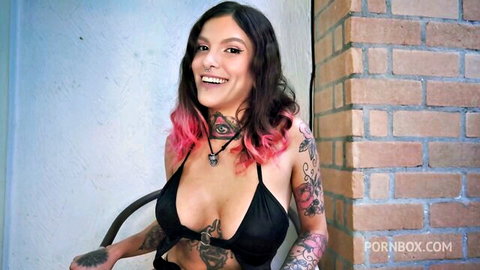 Tattooed Qween Goddess smiling in revealing black top with cleavage