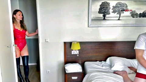 Brunette in red lace bodysuit and thigh-high boots teases man in red briefs on hotel bed