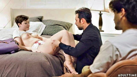 Short-haired brunette with natural tits lies topless on bed, shaved pussy exposed under hiked skirt as man fingers her while cuckold watches.