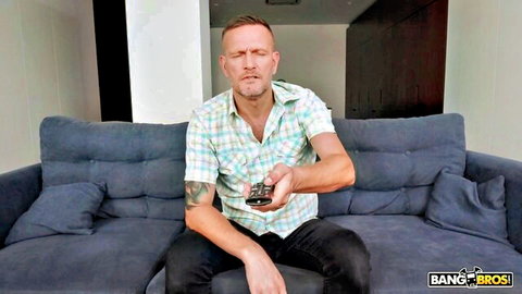 Mature Caucasian man in checkered shirt sits on couch holding remote, looking distressed in emotional Bang Bros scene