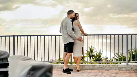 Adria Rae in short dress embraced by man on sunset balcony threesome tease