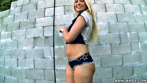 Smiling blonde babe poses with big juicy ass cheeks spilling from tiny star denim shorts against wall