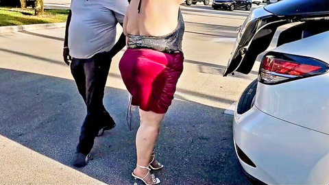 Curvy woman in short red skirt bent over car trunk, exposing ass to man in parking lot