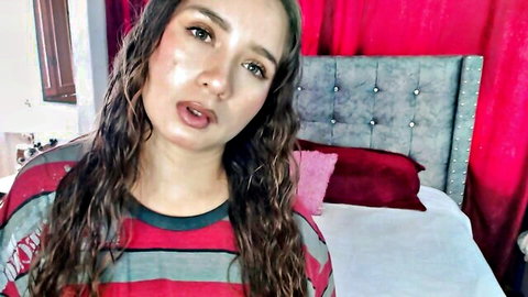 Amateur webcam girl with wavy hair in red striped shirt teasing with open mouth on bed