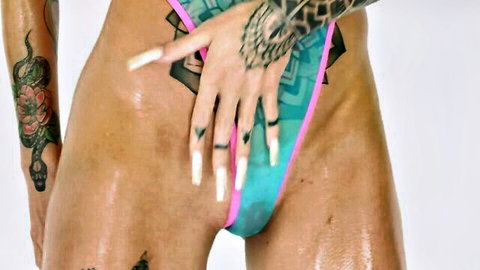 Tattooed woman in skimpy teal thong, oily skin, long white nails, teasing crotch pose
