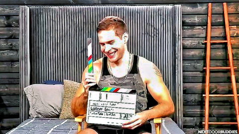 Muscular tattooed guy in tank top smiling holding Next Door Buddies clapperboard on bed set