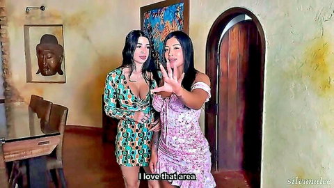 Two busty brunette Latinas in low-cut dresses showing deep cleavage and gesturing seductively indoors