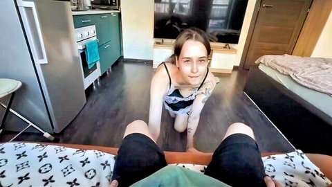 Tattooed brunette on knees in kitchen POV gazing up at cock between black pants legs.