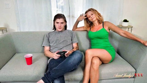 Blonde in tight green dress lounges seductively next to gamer guy on couch