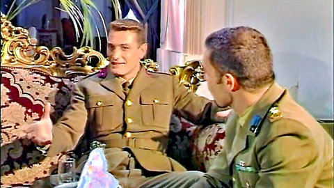Two uniformed soldiers chatting and gesturing on ornate vintage couch, smiling intimately.