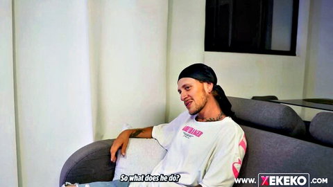 Tattooed guy in white YEKEKO hoodie sits relaxed on gray couch saying 'So what does he?', clothed casual chat.