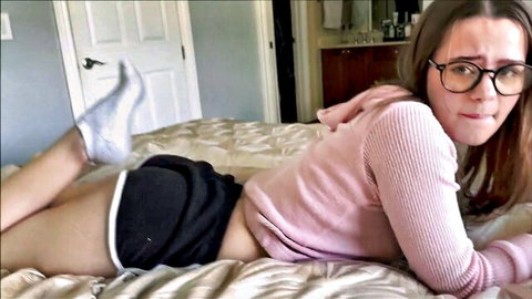 Trinity Olsen in pink hoodie, black shorts, glasses on bed, teasing pose